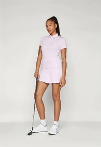 Žena v svetloružovom polo tričku a sukni, ktorá drží golfovú palicu, stojí pred jednoduchým bielym pozadím.