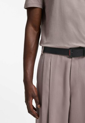 Rechter Arm und Hand einer Person, die ein beigefarbenes Kurzarmhemd und beige plissierte Hosen mit schwarzem Hugo-Gürtel trägt.