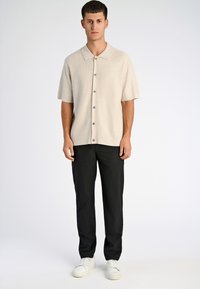 Korte mouw, beige gebreide shirt met een spread collar en knoopsluiting, gecombineerd met zwarte broek en witte sneakers op een lichte achtergrond.