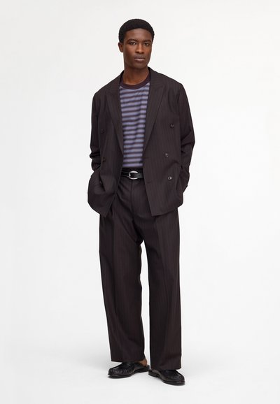 Young man wearing a loose dark pinstripe suit over a striped shirt with black belt and black loafers, standing with hands in pockets.