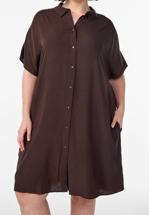 Femme portant une robe ample, à manches courtes, marron foncé, boutonnée avec un col, debout, une main dans la poche, sur fond blanc.