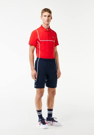 Polo rouge avec des passepoils blancs, shorts navy et chaussures de sport blanches. Présente des logos Lacoste sur le t-shirt et les shorts, chaussettes rayées.