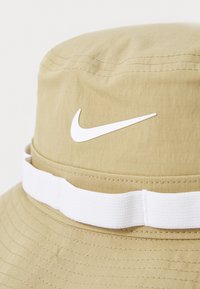 Beige emmerhoed van textiel, met een wit Nike-logo en een witte band rond de onderkant. Stiksels zichtbaar op de kroon.