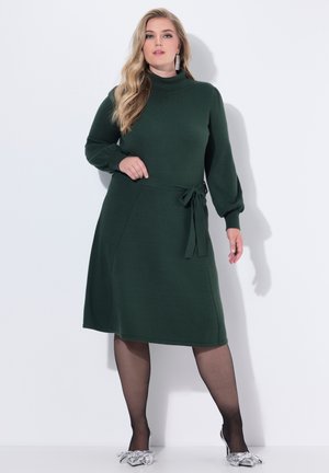 Abito lavorato a maglia verde scuro con scollatura alta, maniche lunghe e cintura legata in vita. Texture liscia, lunghezza al ginocchio, abbinato a scarpe argentate.