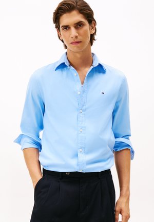 Camicia blu chiaro con bottoni e maniche lunghe, arrotolate ai polsini, e colletto a contrasto. Presenta un piccolo logo sul petto e una texture liscia.