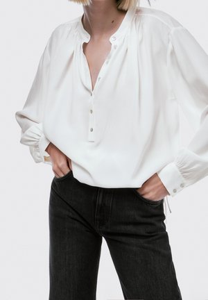 Femme portant un chemisier blanc ample boutonné avec des détails froncés et un jean noir taille haute, les mains dans les poches.