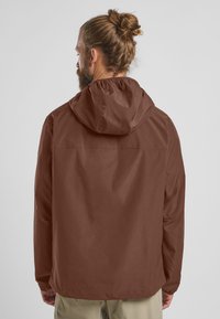 Bruine waterdichte jas met capuchon, voorzien van een gladde textuur en elastische manchetten. Recht gesneden ontwerp met een rugpas-detail.