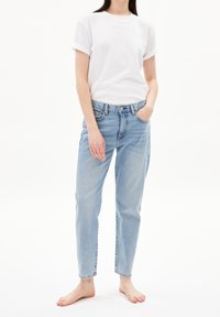 Femme portant un t-shirt blanc à manches retroussées et un jean bleu clair, debout pieds nus avec une main dans la poche, sur un fond blanc.