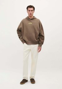 Brązowy oversize'owy sweter z kieszenią z przodu i kapturem, z logo. W połączeniu z kremowymi spodniami i brązowymi trampkami.