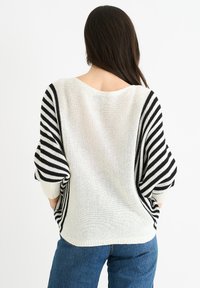 Sweter dzianinowy o luźnym, oversize'owym kroju, z białym korpusem i czarnymi poziomymi paskami na rękawach. Teksturowana tkanina.