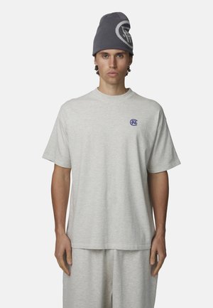 Un giovane indossa una maglietta oversize grigio chiaro e pantaloni abbinati con un berretto grigio scuro caratterizzato da un logo rotondo.