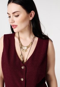 Bordeauxrood textuurtop met een diepe v-hals, voorzien van knopen in goudkleur en gelaagde gouden kettingen met verschillende metalen details.