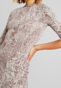 Polven pituinen, vartalonmyötäinen mekko, jossa on beige ja ruskea käärmeennahkakuosi. Mekko on valmistettu rakenteellisesta materiaalista, jossa on korkea kaulus ja puolihihat.