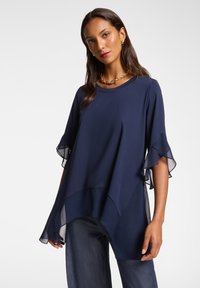 Top blu navy con maniche corta a strati trasparenti, scollatura rotonda e vestibilità comoda. Abbinato a jeans in denim scuro per un look casual.