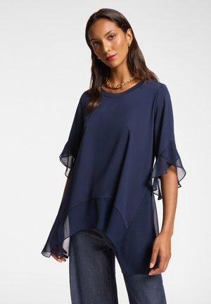 Navy blue top with short, layered sheer sleeves, round neckline, and a relaxed fit. Paired with dark denim jeans for a casual look.