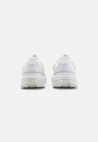 White sneakers with a mesh upper and rubber sole, featuring a curved shape, prominent heel cushioning, and minimalistic design.