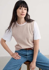 Világos bézs kötött mellény egy fehér póló felett, kék farmernadrággal párosítva. A mellény bordázott nyakkivágással és karöltőkkel rendelkezik, lazán illeszkedik.