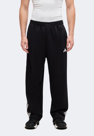 Osoba ubrana w czarne spodnie dresowe Adidas z białymi paskami i logo, zestawione z czarno-białymi sneakersami i białym dopasowanym topem.