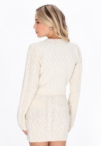 Sweter sukienka w kolorze kremowym, dziergany, z długimi rękawami, teksturowanymi wzorami w warkocze, dopasowaną talią i subtelnym połyskiem. Posiada okrągły dekolt i długość mini.