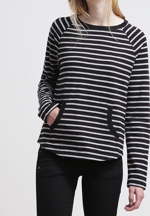 Femme portant un haut à manches longues rayé noir et blanc avec une poche avant, associée à un pantalon noir, se tenant devant un fond uni.