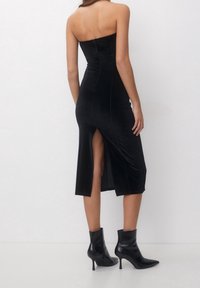 Vestido midi negro sin tirantes con un diseño ajustado, abertura lateral y cierre de cremallera en la parte posterior. Textura suave y silueta elegante.