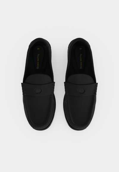 Ζευγάρι μαύρα loafers χωρίς κορδόνια με ραμμένες λεπτομέρειες και μικρό διακοσμητικό έμβλημα στο πάνω μέρος, μπροστά σε λευκό φόντο.