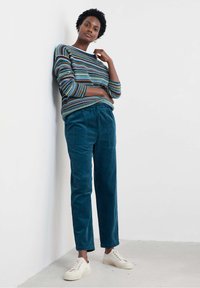 Maglione a righe in multicolore; pantaloni in velluto a coste teal con vestibilità rilassata; scarpe bianche con stringhe; sfondo neutro.