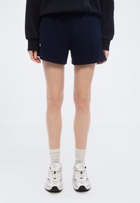 Άτομο που στέκεται φορώντας ναυτικά σορτς, λευκές κάλτσες, λευκά αθλητικά παπούτσια και μαύρο μακρυμάνικο πάνω μέρος μπροστά σε απλό λευκό φόντο.