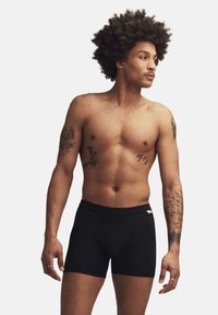 Svarta boxershorts med en mjuk elastisk midjeband, åtsittande design och slät textur, bärs av en modell med tatueringar och lockigt hår.