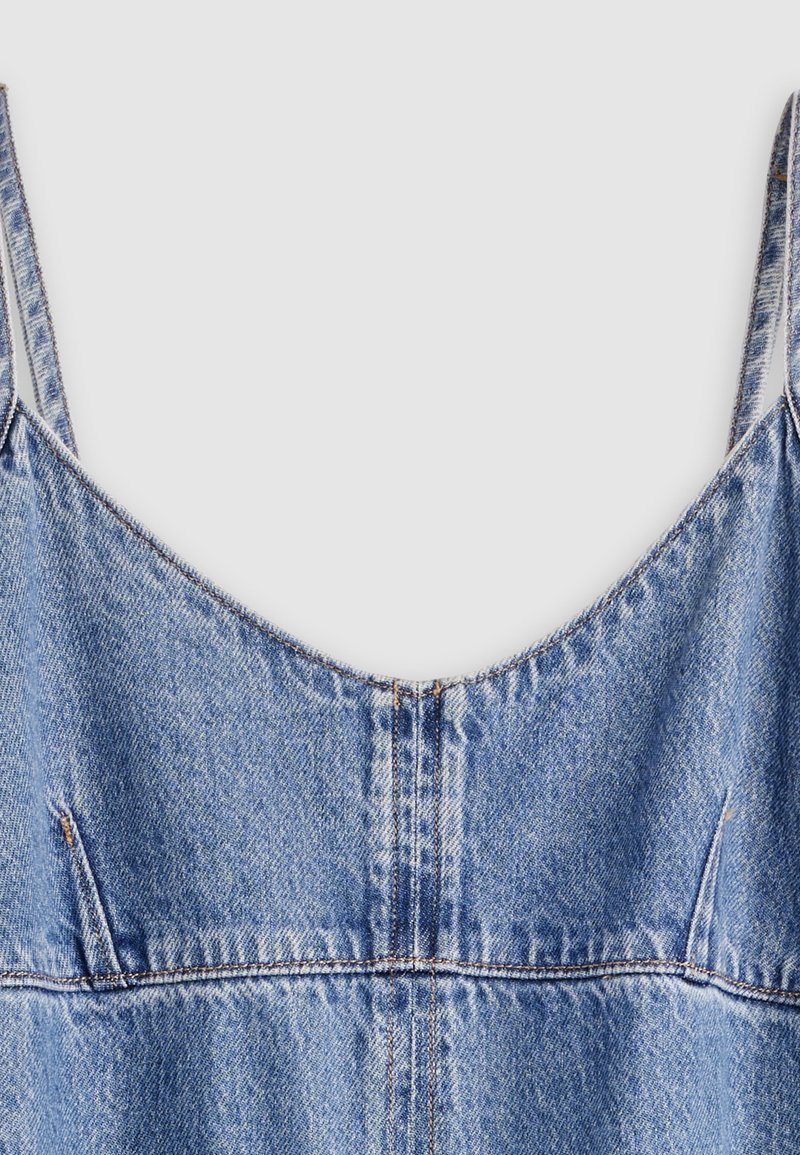 Hellblaues Denim-Kleid mit dünnen Schulterriemen und sichtbaren Nahtdetails vor einem einfarbigen Hintergrund.
