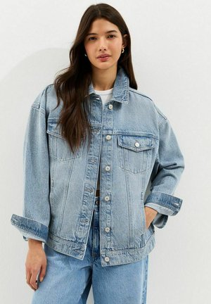 Jonge vrouw met lang bruin haar, gekleed in een oversized lichtblauwe spijkerjasje en bijpassende spijkerbroek, staand tegen een effen witte achtergrond.