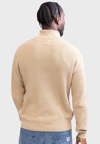 Beige strikkegenser med høy krage, med teksturert ribbing og en tettsittende nederkant, sett bakfra.