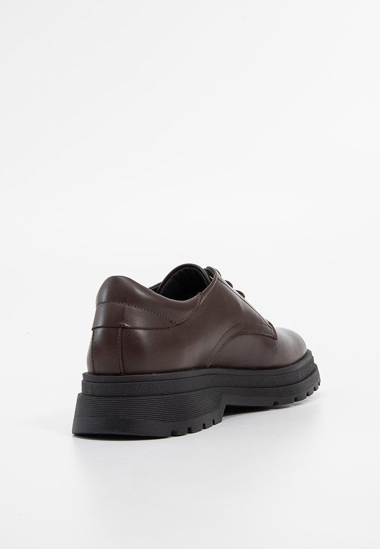 Brązowe skórzane buty o zaokrąglonym kształcie, niskim obcasie i teksturowanej czarnej gumowej podeszwie. Posiadają gładkie wykończenie i minimalne detale szycia.