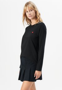 Vrouw in een zwart langemouwenshirt met een klein rood hartdetail en een zwarte geplooide minirok, staande tegen een witte achtergrond.