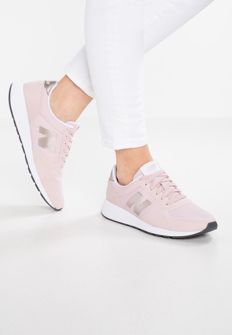 Personne portant des baskets New Balance rose clair et un pantalon blanc, debout sur un sol blanc et lisse.