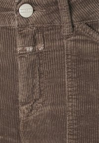 Pantalones de pana marrones con textura de costillas verticales, que presentan un botón metálico etiquetado como "CLOSED" y un bolsillo lateral con detalles de costura.