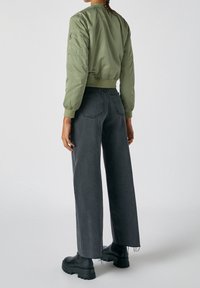 Personne portant une veste bomber vert olive, un jean noir large taille haute et des bottes noires épaisses, debout de dos.