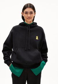 Felpa nera con cappuccio e tasca frontale, decorata con un piccolo emblema a nastro giallo. Sottostante una camicia verde con colletto.