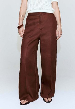 Persona con pantalones marrones de pernera ancha, top blanco de punto sin mangas, sandalias negras y pulseras, de pie frente a un fondo liso y claro.