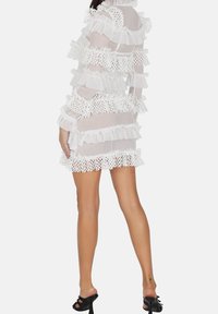 Robe blanche transparente avec des volants en couches et des détails en dentelle. Elle possède des manches longues et une silhouette ajustée, se terminant au milieu de la cuisse.