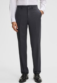 Mężczyzna w ciemnoszarych, wzorzystych eleganckich spodniach, czarnych eleganckich butach i białej koszuli z długim rękawem, stoi z jedną ręką w kieszeni.