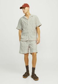 Camisa de manga corta texturizada en gris claro y pantalones cortos a juego, combinados con calcetines marrón oscuro y sandalias. Gorra roja con un logo en la parte delantera.