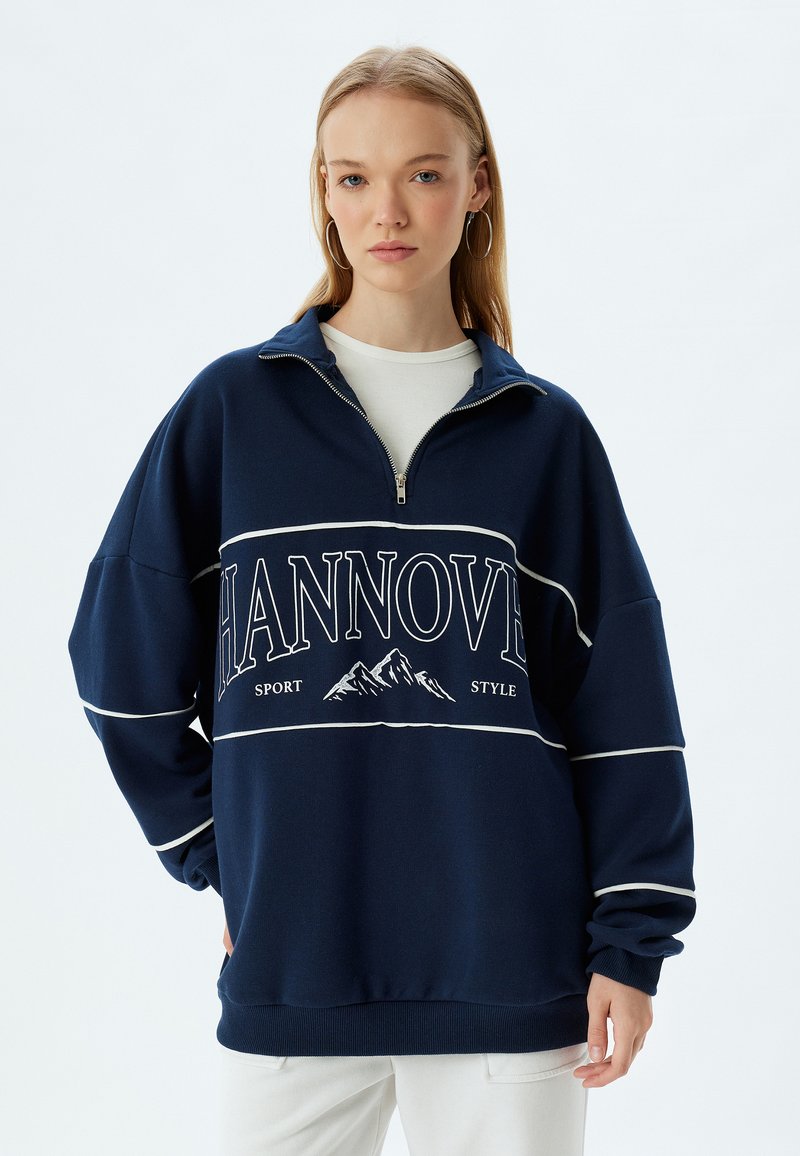 Sudadera navy con cuello de media cremallera, que presenta un diseño gráfico blanco con "HANNOVER" e ilustración de montañas en la parte delantera.
