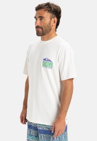 Camiseta blanca de manga corta con un colorido logo de Quiksilver en el pecho. También incluye pantalones cortos azules con patrones de diseños geométricos.