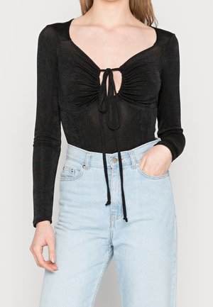 Woman wearing a black long-sleeve top with front tie detail paired with light blue high-waisted jeans, one hand in pocket.