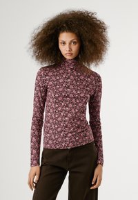 Langsleeve turtleneck-top in donkerbruin met een roze bloemenpatroon. Zachte, rekbare stof met een getailleerde pasvorm. Zwarte broek zichtbaar.