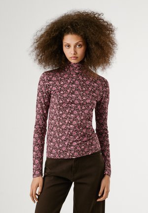 Langsleeve turtleneck-top in donkerbruin met een roze bloemenpatroon. Zachte, rekbare stof met een getailleerde pasvorm. Zwarte broek zichtbaar.