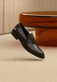 Mocassins en cuir noir à finition lisse, avec une bande texturée et un bout arrondi, posés sur un fond beige doux.