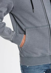 Felpa grigia con zip e finitura texturizzata, tasche frontali, polsini e orlo a coste. Presenta un cordino nero per la regolazione.