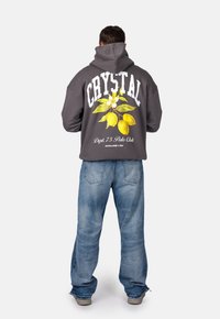 Felpa con cappuccio grigia caratterizzata da una grafica floreale di limone e lettering bianco sulla schiena, abbinata a jeans baggy blu chiaro.